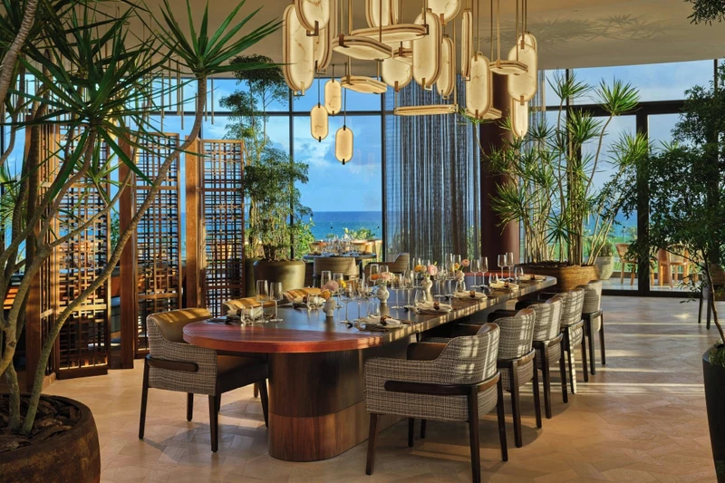 A spacious dining area with a long table, multiple chairs, hanging pendant lights, and large windows offering a view of the ocean.