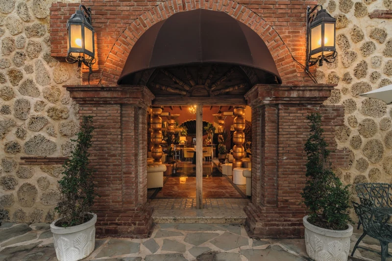 A brick entrance with two lanterns, a stone wall, and a doorway leading to an interior with furniture.
