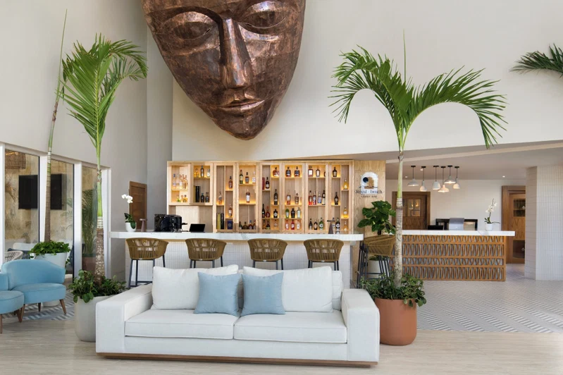 A spacious lobby with a reception desk, a bar area with shelves of bottles, a white sofa with blue cushions, and two large potted plants.