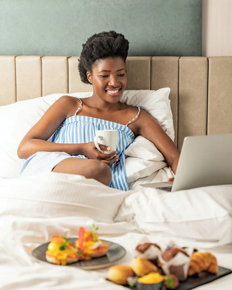 A person in a bed with a laptop, a cup, and breakfast items on a tray.