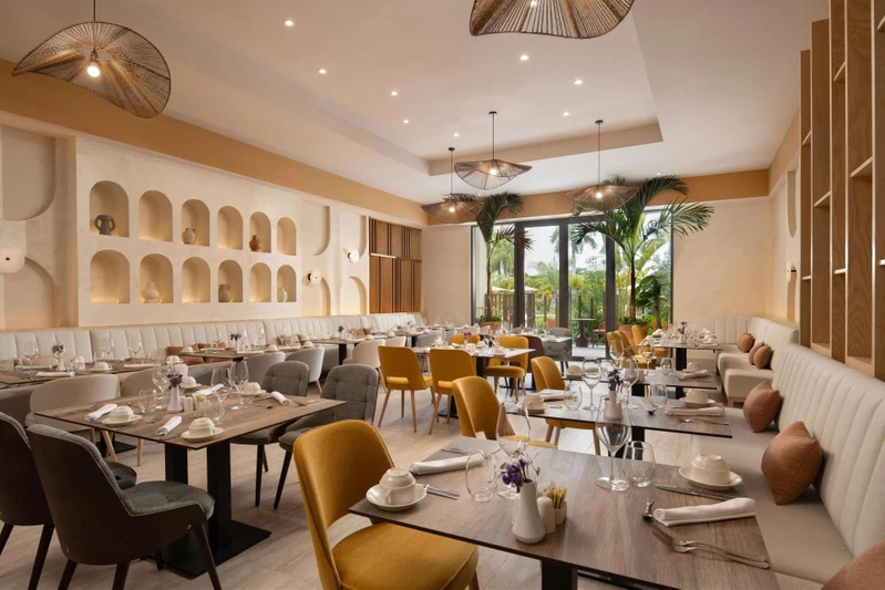 A modern restaurant with multiple dining tables, comfortable seating, and a view of greenery outside.