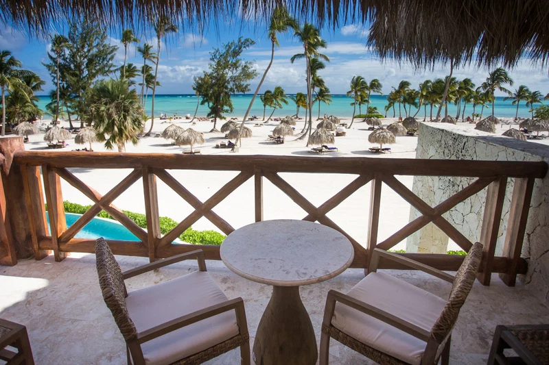 A beachside patio with a round table and wicker chairs, overlooking a sandy beach with palm trees and a clear blue sea.