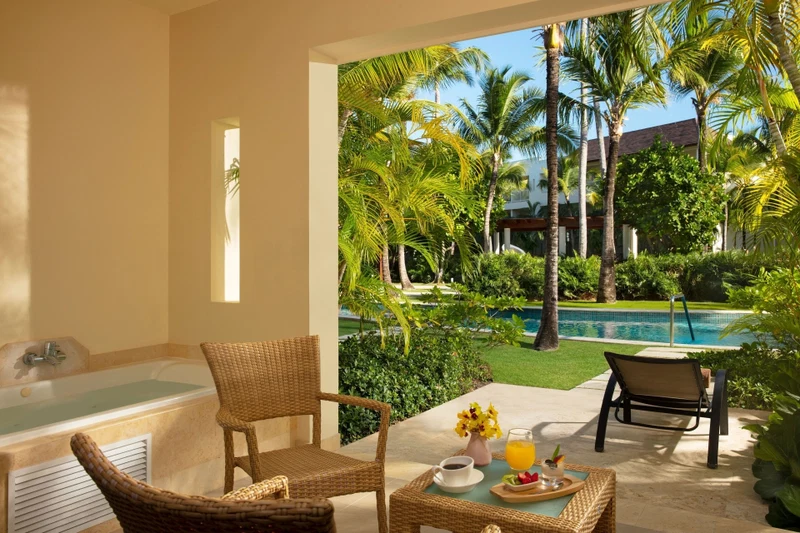 A room with a bathtub, wicker chairs, a table with breakfast items, and a view of a pool and palm trees.
