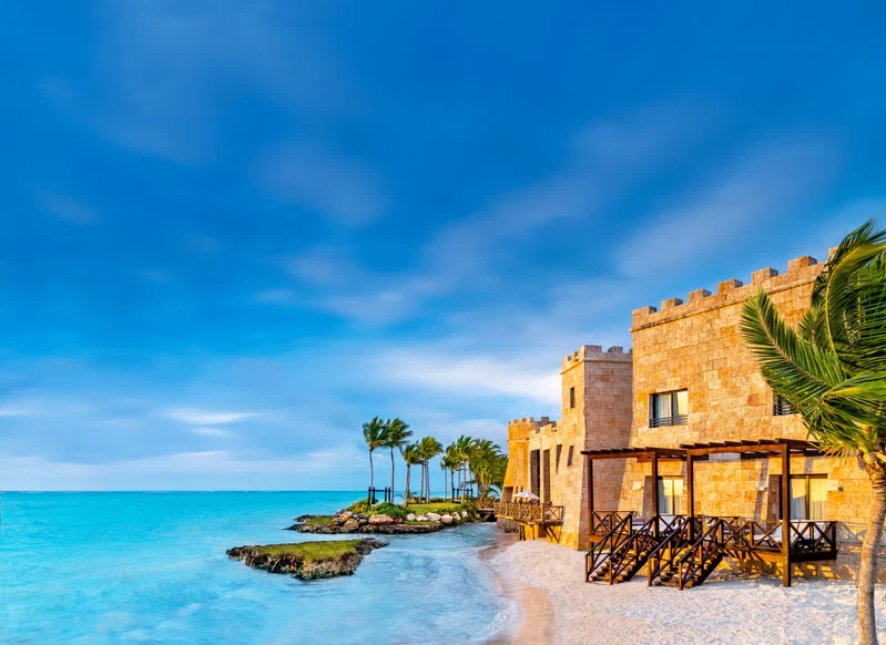 A beachfront hotel with a stone facade, palm trees, and a clear blue sky.