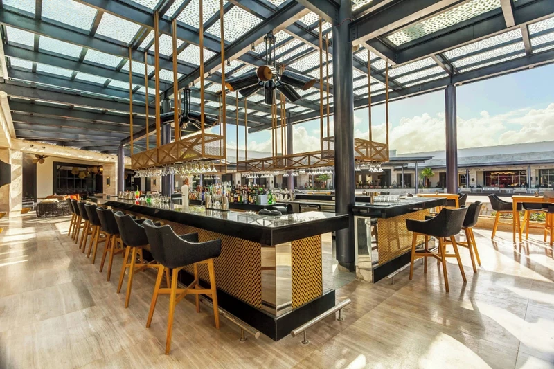 A spacious bar area with high ceilings and a modern design, featuring a long counter with stools and a variety of bottles.