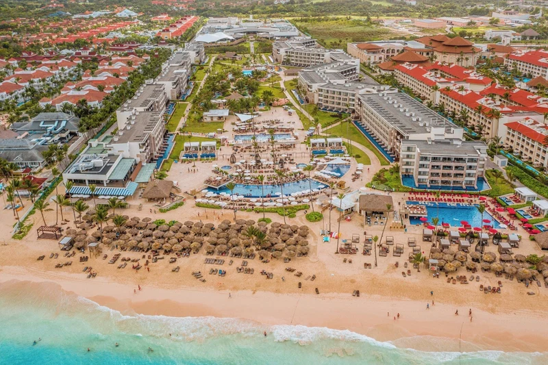 An aerial view of a resort with multiple swimming pools, a beach area with sun loungers, and a variety of buildings with red roofs.