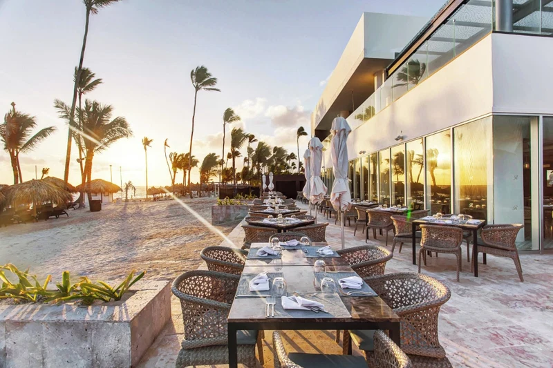 An outdoor dining area with tables and chairs, palm trees, and a modern building with glass windows.