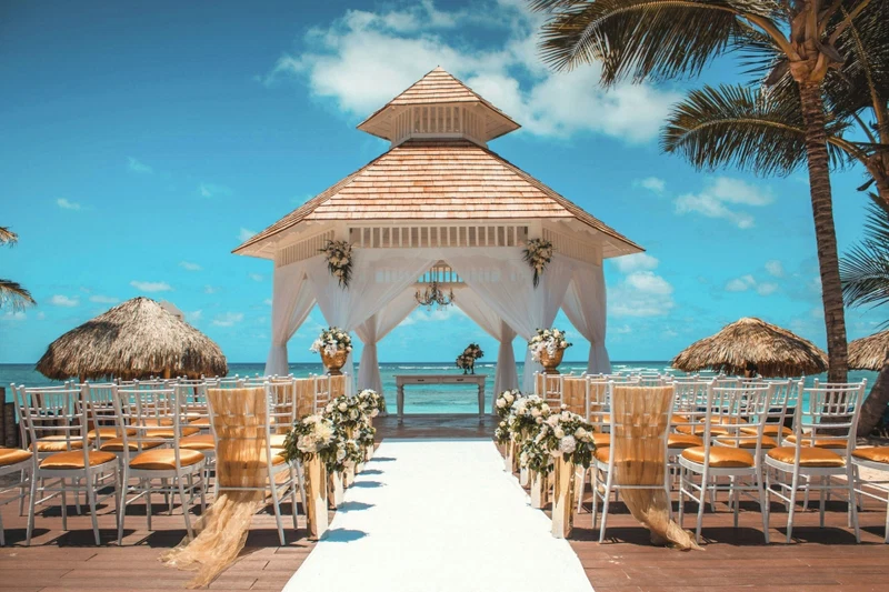 A beach wedding setup with a wooden gazebo, white chairs, and a white aisle.