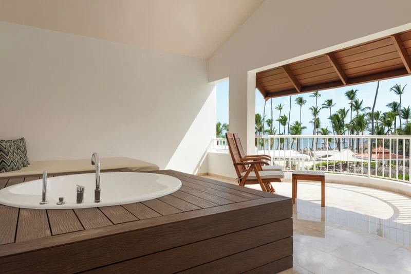 A modern bathroom with a wooden vanity, a large bathtub, and a balcony with a view of palm trees and a pool.