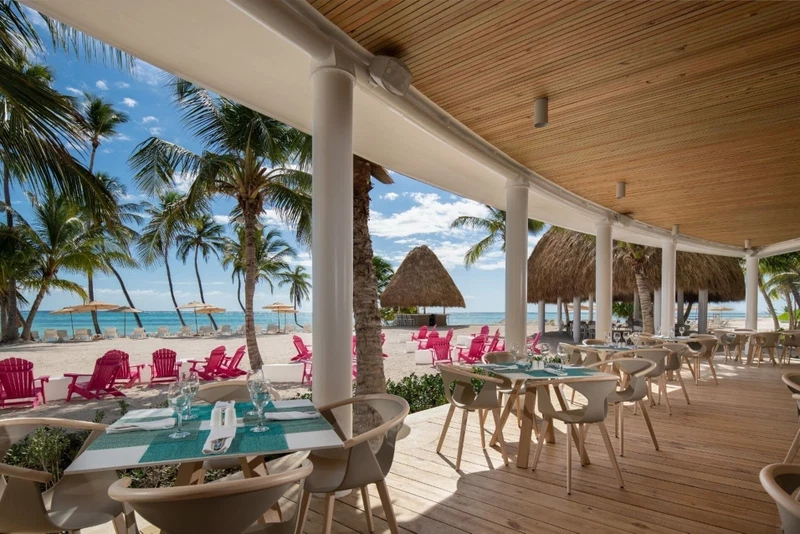 A beachside restaurant with outdoor seating, palm trees, and a view of the ocean.