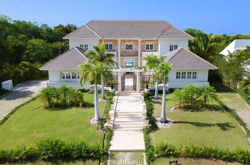 A large two-story house with a central entrance, surrounded by a well-maintained lawn and palm trees.