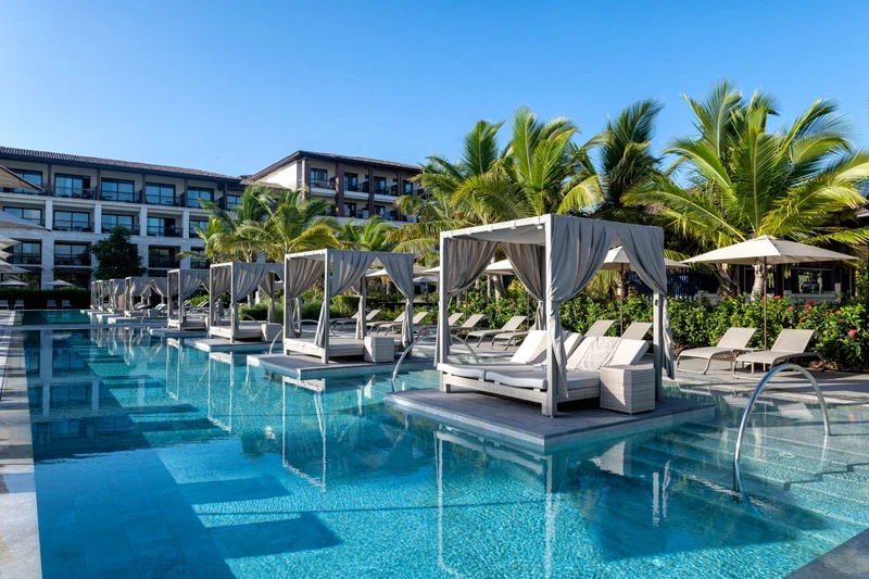 A pool area with lounge chairs and umbrellas, surrounded by palm trees and a building in the background.