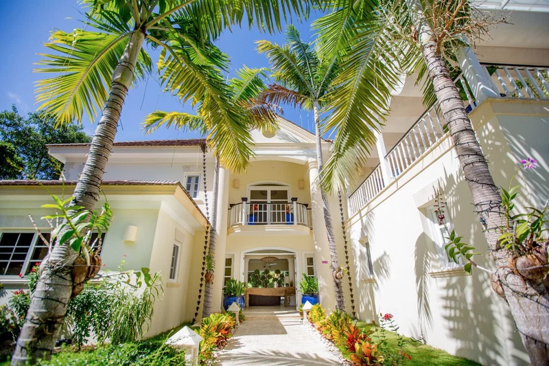 A two-story building with a central entrance, surrounded by palm trees and potted plants.