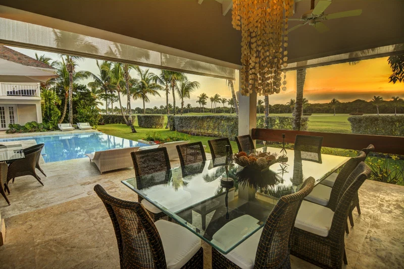 A glass-top dining table with wicker chairs, a pool, and a view of a golf course at sunset.