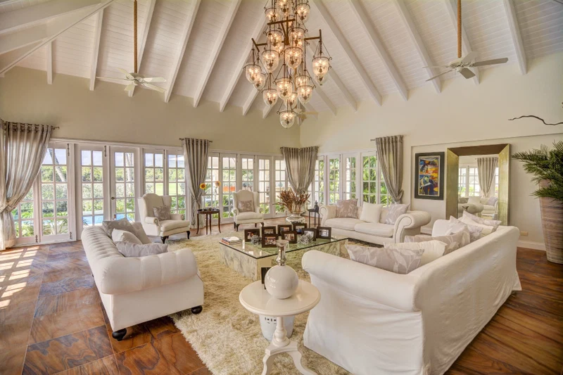 A spacious living room with white furniture, large windows, and a chandelier.