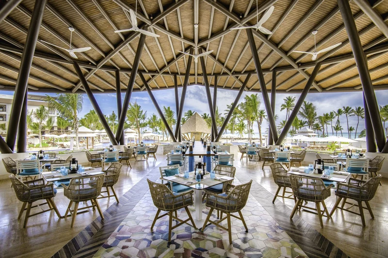A spacious outdoor dining area with multiple tables and chairs, a central island with a bar, and a view of palm trees and the ocean.