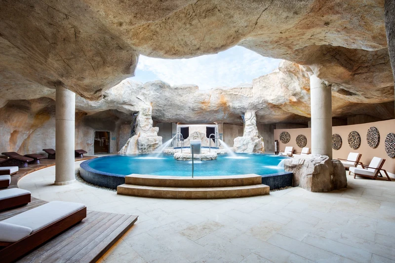A spa area with a hot tub, lounge chairs, and a pool under a rock formation.