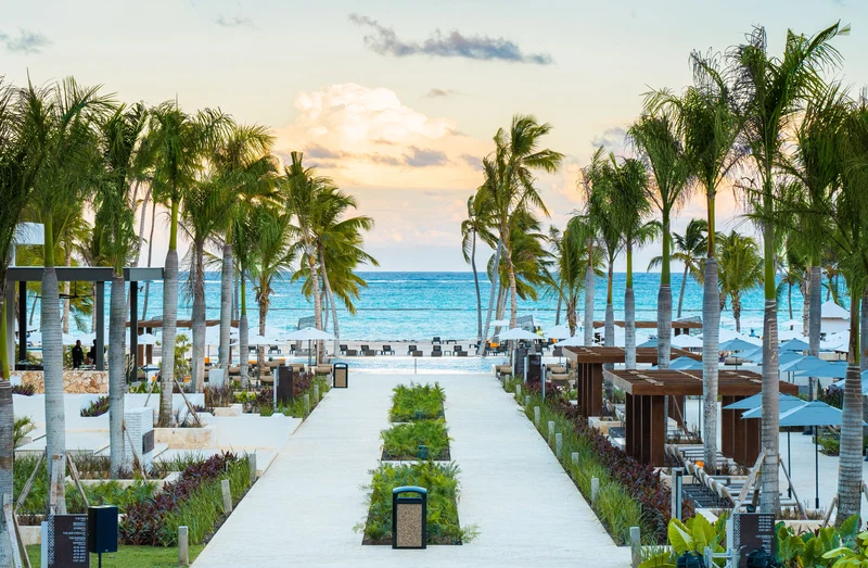 A beachfront resort with a pathway lined with palm trees and beach umbrellas, leading to the ocean.