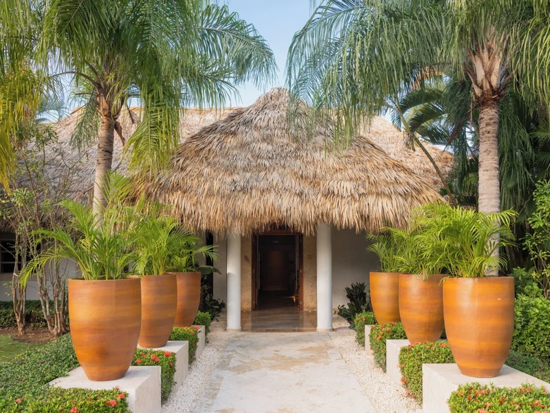 A tropical resort entrance with a thatched roof, palm trees, and large potted plants.