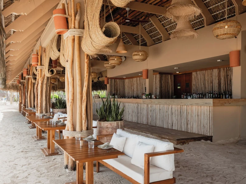 A beachside bar with wooden tables and benches, a thatched roof, and a variety of hanging lights.
