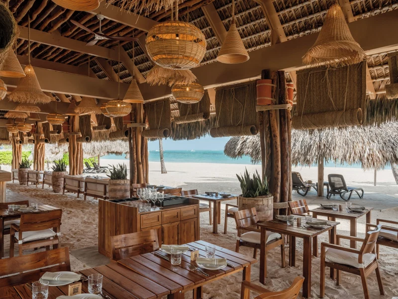A beachside restaurant with wooden tables and chairs, thatched umbrellas, and a view of the ocean.
