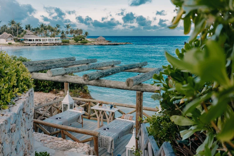 A wooden deck with seating and a table, overlooking a turquoise sea with palm trees and a thatched-roof structure in the background.