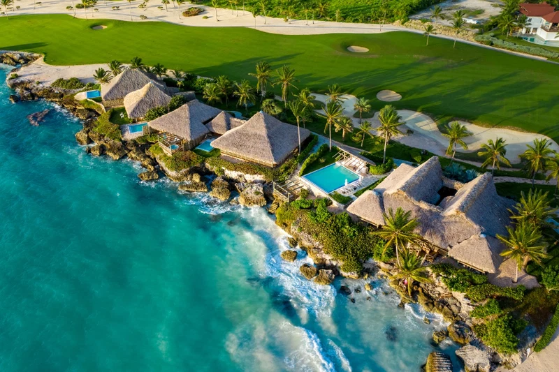 An aerial view of a coastal resort with thatched-roof villas, a swimming pool, and a golf course.