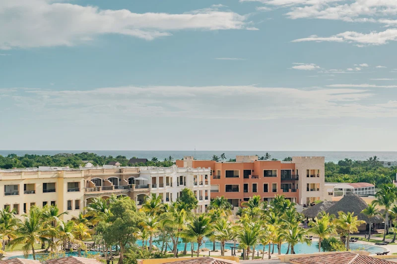A resort with multiple buildings, a pool area with palm trees, and a view of the ocean.