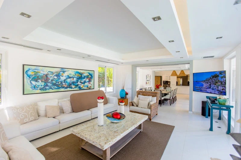 A modern living room with a white sofa, a coffee table, and a TV. There is a painting on the wall and a dining area in the background.