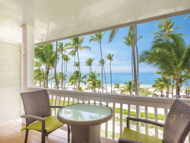 A balcony with wicker chairs and a glass table overlooking a beach with palm trees and the ocean.