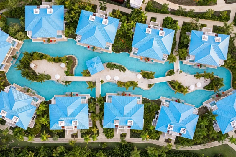 An aerial view of a resort with multiple blue-roofed villas arranged around a central swimming pool, surrounded by lush greenery and palm trees.