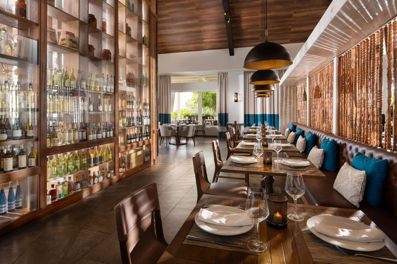 A restaurant with a long dining area, wooden tables, and a bar with glass-fronted shelves of bottles.