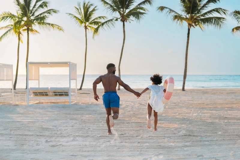 A beach scene with a man and a child holding hands, palm trees, and a beach umbrella.