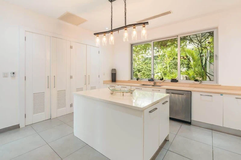 A modern kitchen with white cabinets, a central island, a stainless steel dishwasher, and a large window with a view of greenery.