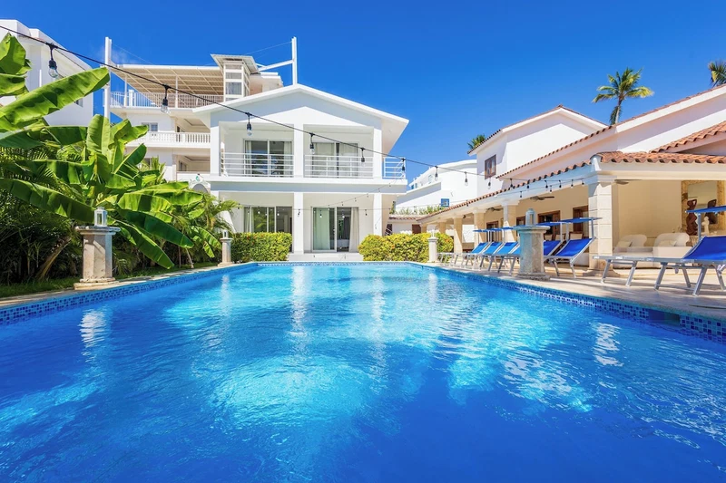 A swimming pool with a clear blue water, surrounded by white buildings with balconies, and a few palm trees.