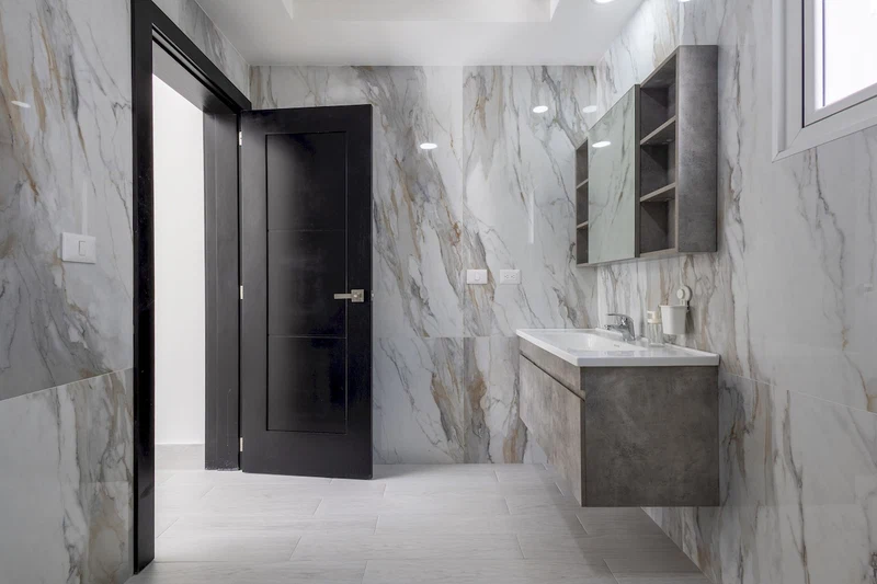 A modern bathroom with marble walls, a dark door, and a built-in sink with a mirror above it.