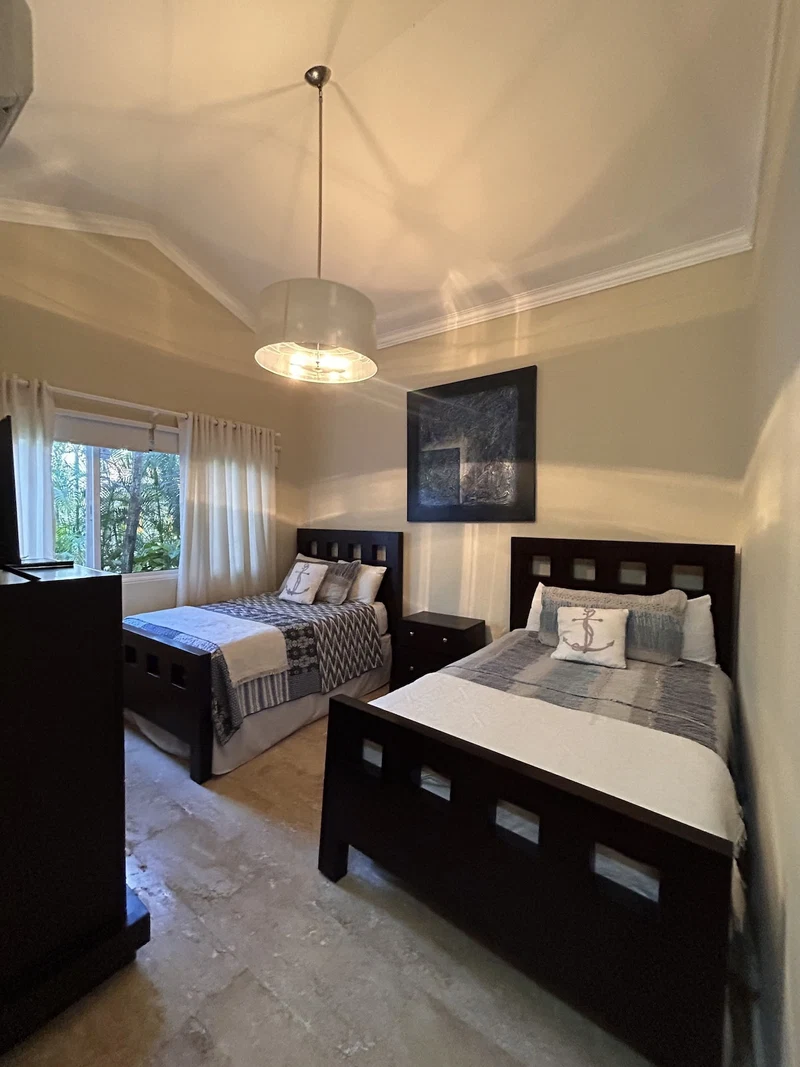 A bedroom with two beds, a ceiling light, a window with curtains, and a painting on the wall.