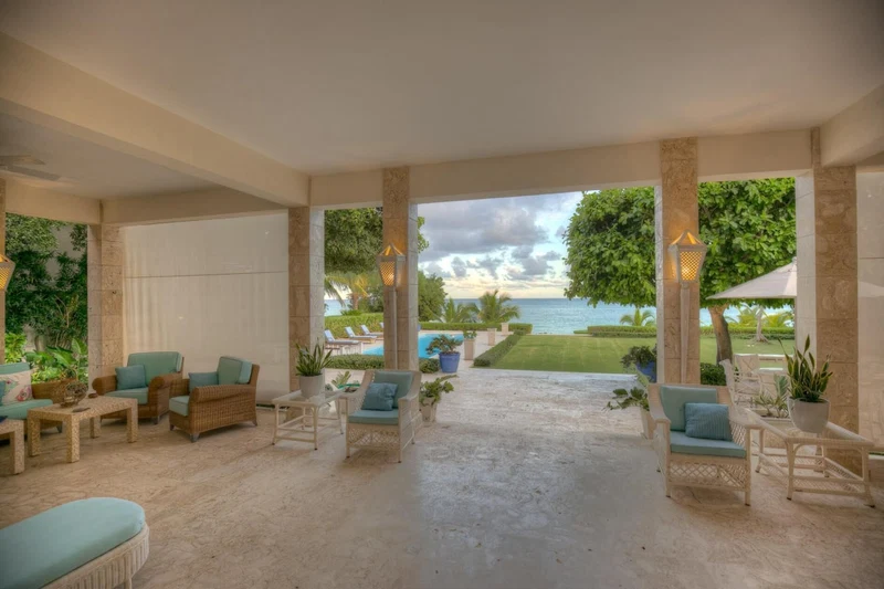 A spacious indoor area with a view of the ocean, furnished with wicker chairs and tables, and surrounded by greenery.