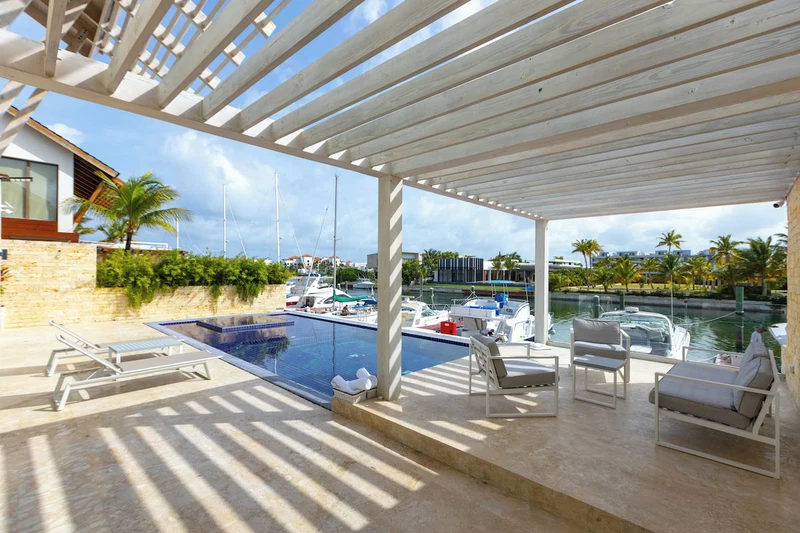 A covered outdoor pool area with lounge chairs, a small pool, and a view of boats and palm trees.
