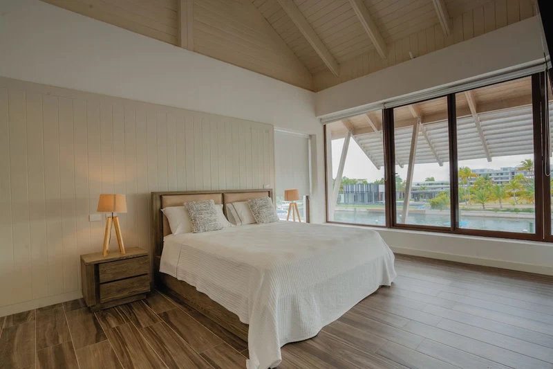 A bedroom with a large bed, wooden floor, and a view of a body of water through large windows.