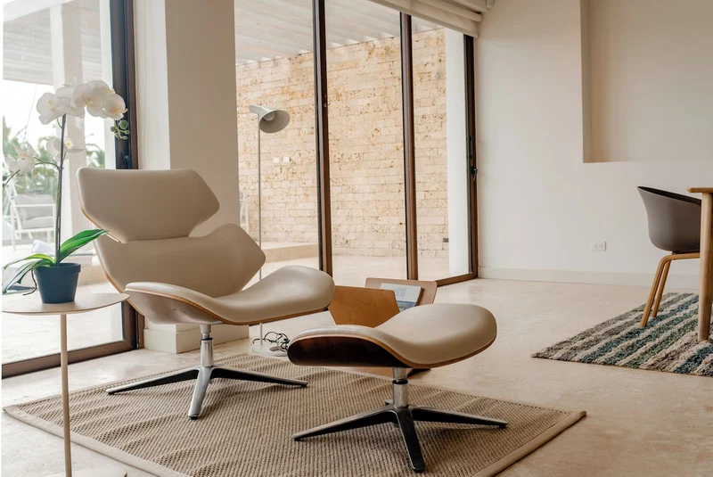 A modern living room with a white leather chair, a small side table with a potted plant, and a large window with a view of a brick wall.