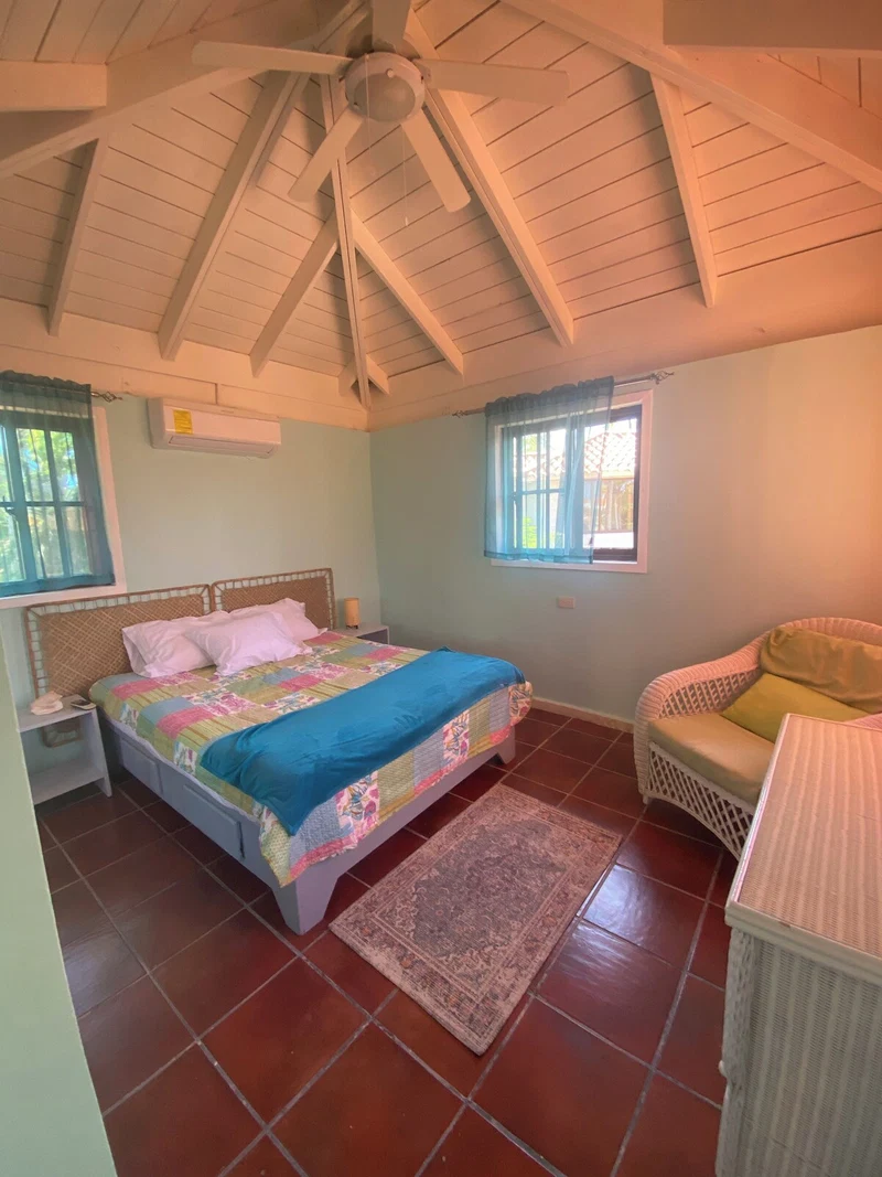 A bedroom with a bed, a nightstand, a wicker chair, a window with curtains, and a ceiling fan.