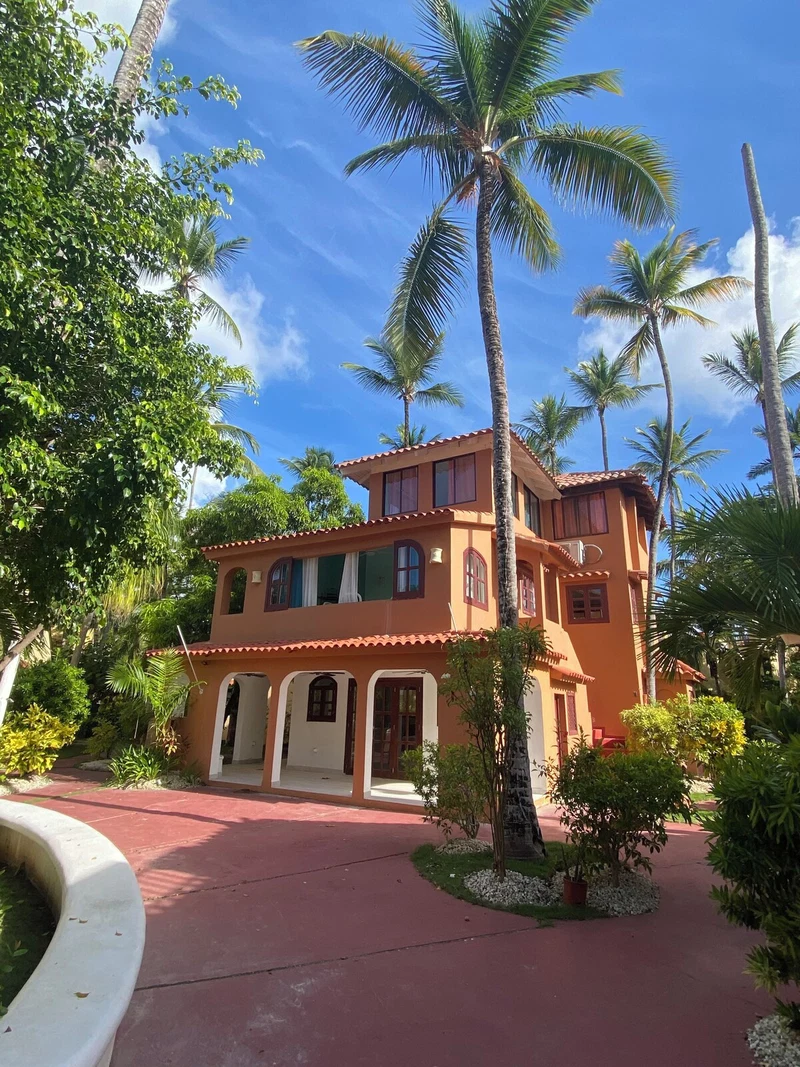 A two-story building with a red-tiled roof, surrounded by palm trees and lush greenery.