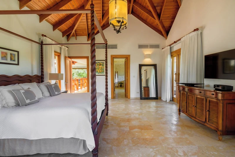 A bedroom with a four-poster bed, wooden ceiling beams, a chandelier, a large mirror, and a television on a wooden cabinet.