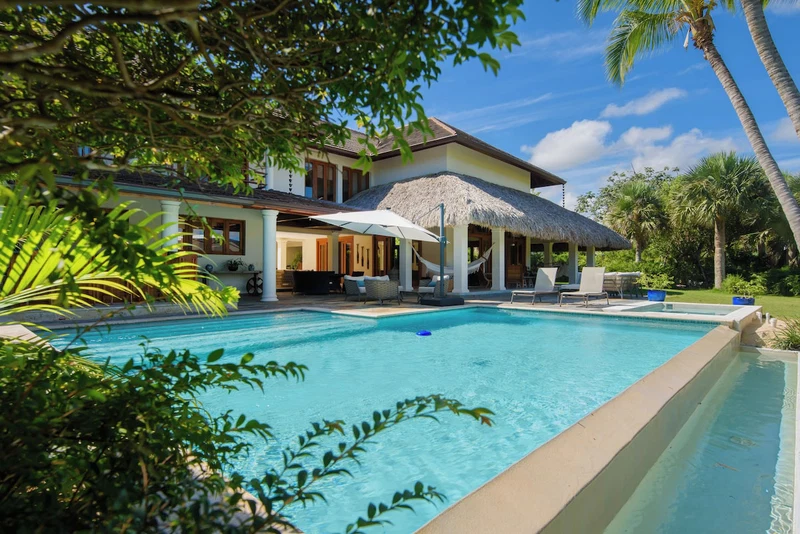 A pool area with a thatched-roof structure, lounge chairs, and palm trees.