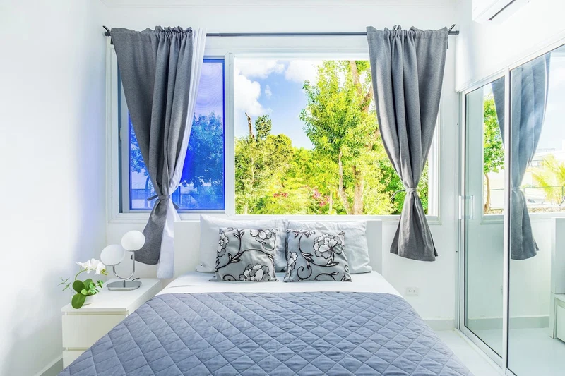 A bedroom with a bed, a nightstand, a window with curtains, and a view of greenery outside.