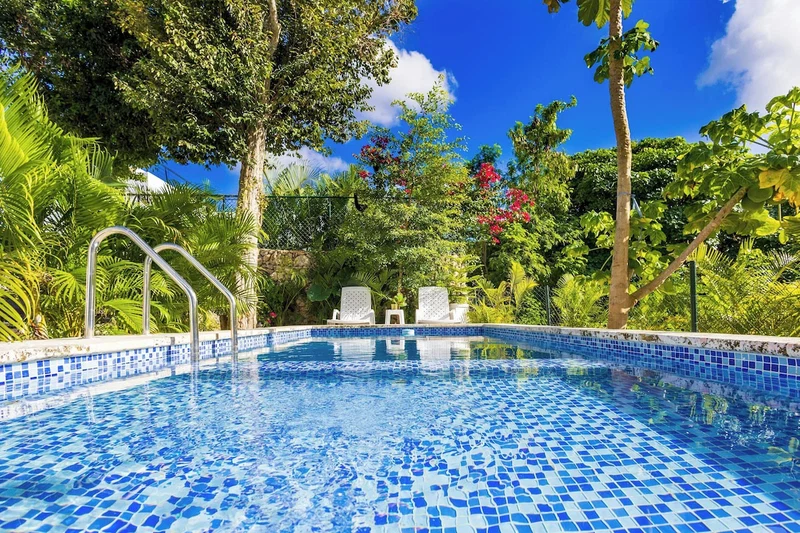 A swimming pool with blue mosaic tiles, surrounded by lush greenery and white lounge chairs.