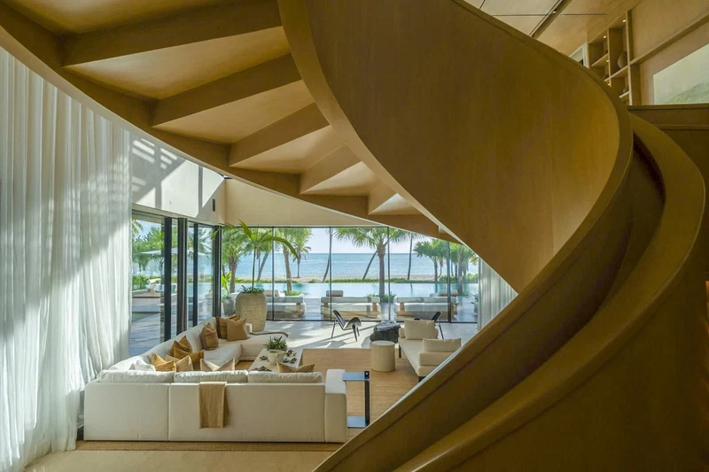 A modern interior with a curved wooden staircase, a white sofa set, and a view of the ocean.