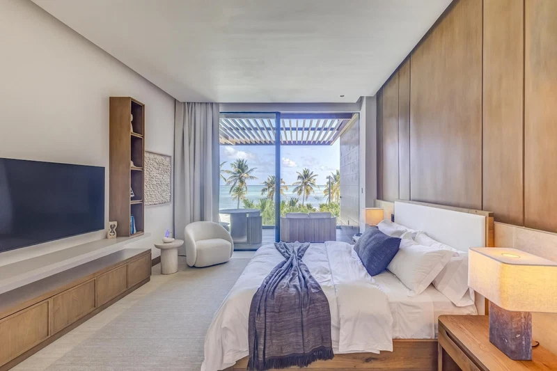 A modern hotel room with a large bed, a TV, a seating area with a view of the ocean, and a balcony with palm trees.