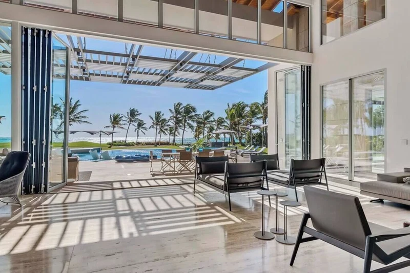 A modern interior with a large glass door leading to a pool area with palm trees and a clear view of the ocean.